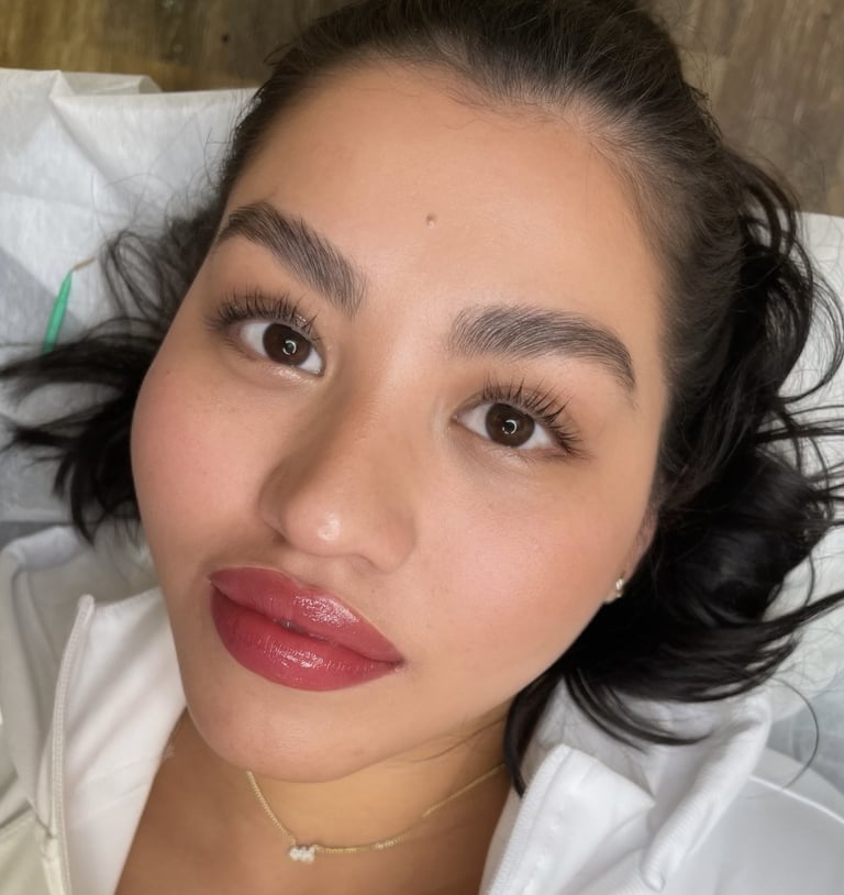 Close-up portrait of a woman with dark hair, defined eyebrows, pink-mauve lipstick, and a gold necklace against a white pillow