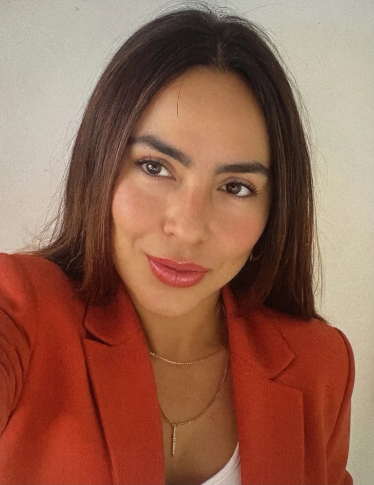 Professional headshot of a woman with long dark hair wearing a red blazer and gold necklace against a neutral background